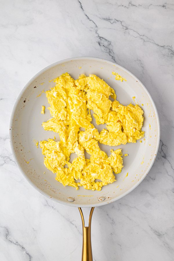 Scrambled eggs cooking in a white nonstick frying pan with a gold handle on a light marble countertop.