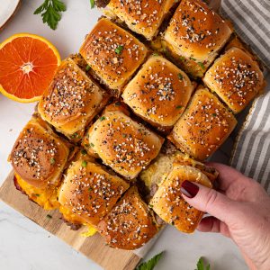 A hand lifts a slider from a tray of baked sandwich sliders topped with sesame seeds, next to half an orange and a striped napkin on a white surface.