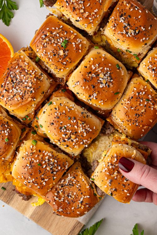A hand lifts a slider from a tray of baked sandwich sliders topped with sesame seeds, next to half an orange and a striped napkin on a white surface.
