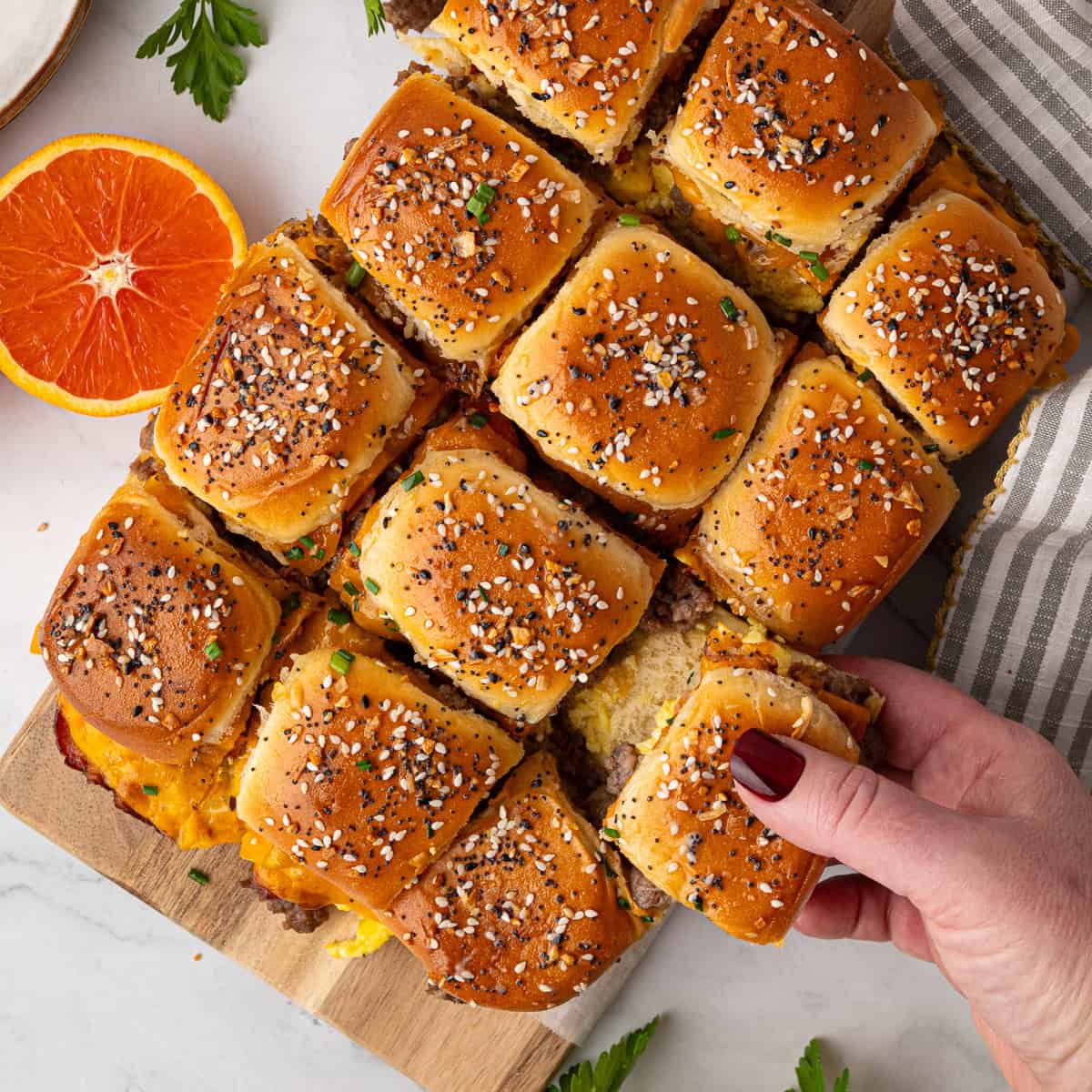 A hand lifts a slider from a tray of baked sandwich sliders topped with sesame seeds, next to half an orange and a striped napkin on a white surface.