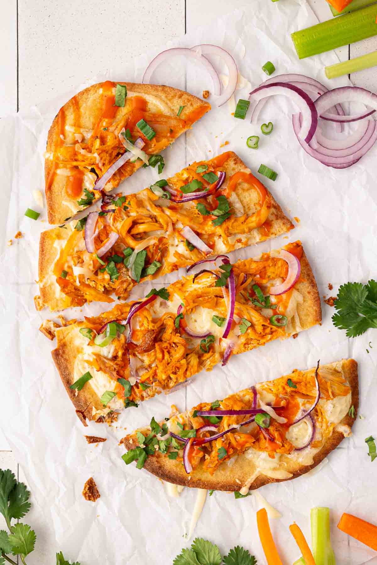A flatbread pizza topped with shredded chicken, melted cheese, red onion slices, green onions, and cilantro, on parchment paper with celery, carrot sticks, and fresh herbs nearby.