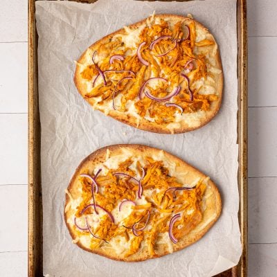 Two flatbreads topped with shredded chicken, cheese, and red onion on a baking sheet lined with parchment paper.