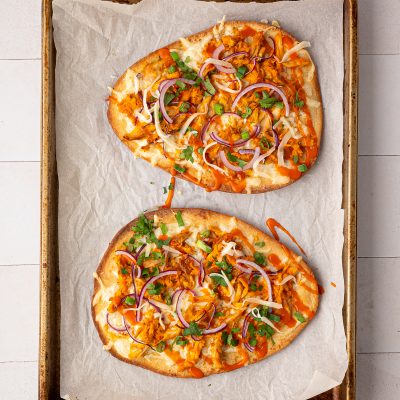 Two flatbread pizzas topped with shredded chicken, red onion, chopped cilantro, and cheese on a parchment-lined baking sheet.