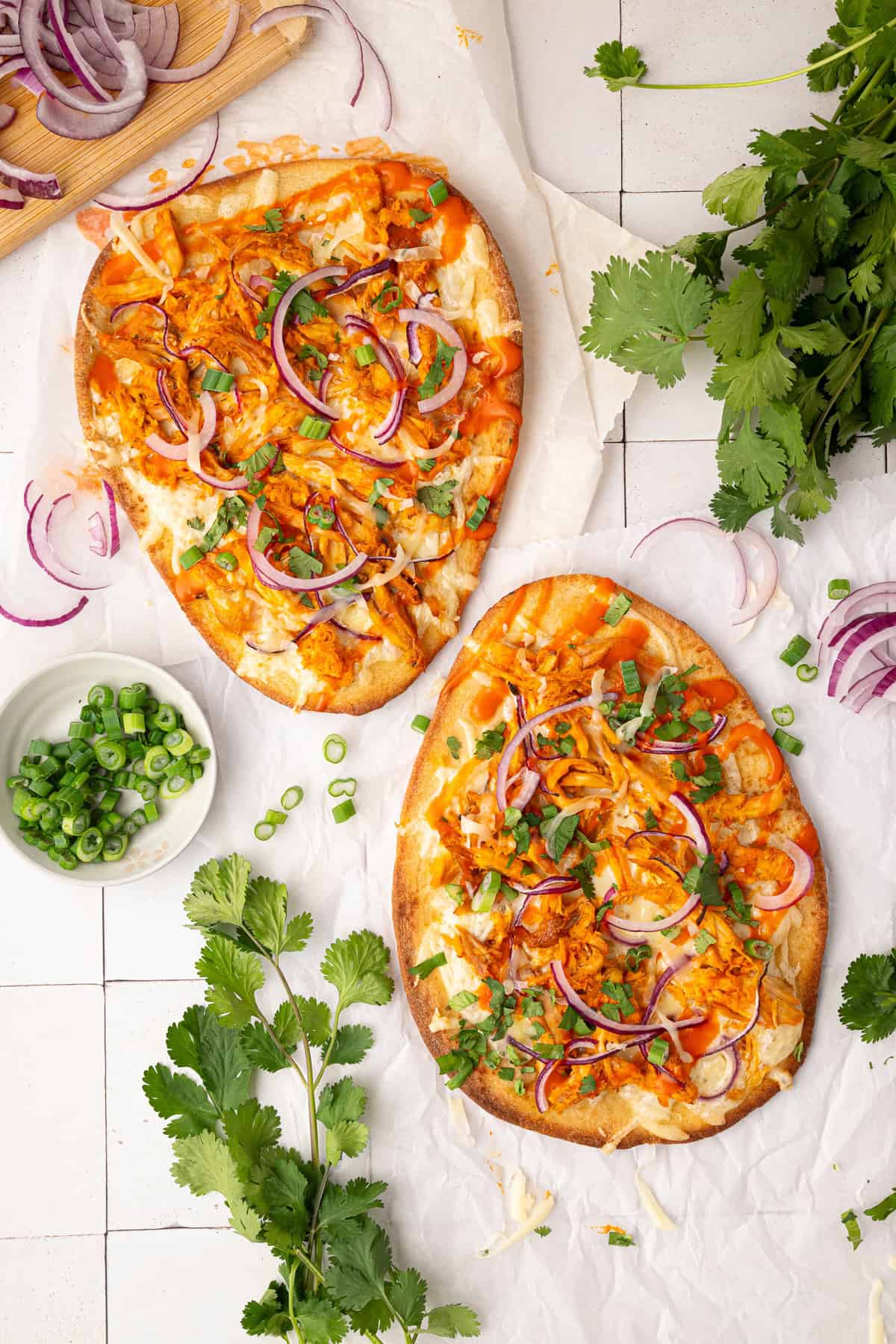 Two flatbread pizzas topped with chicken, red onion, cheese, and chopped cilantro sit on parchment paper, surrounded by fresh herbs and sliced green onions.