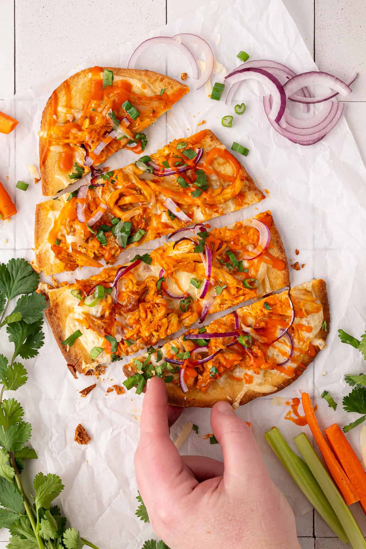 A hand reaches for a slice of flatbread topped with shredded chicken, red onions, green onions, and sauce, on parchment paper with fresh herbs and vegetable sticks nearby.