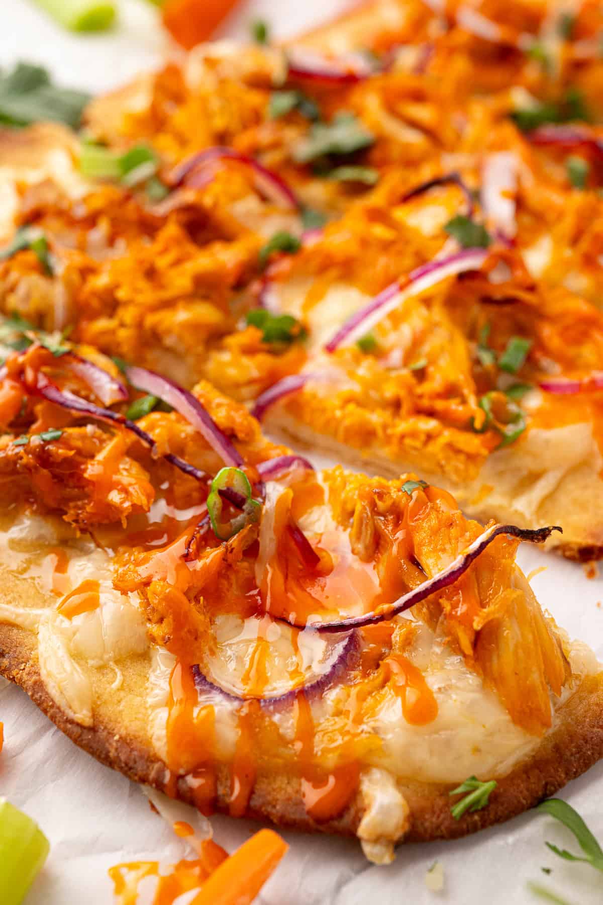 Close-up of a flatbread topped with shredded buffalo chicken, melted cheese, red onion slices, chopped green herbs, and a drizzle of orange sauce.