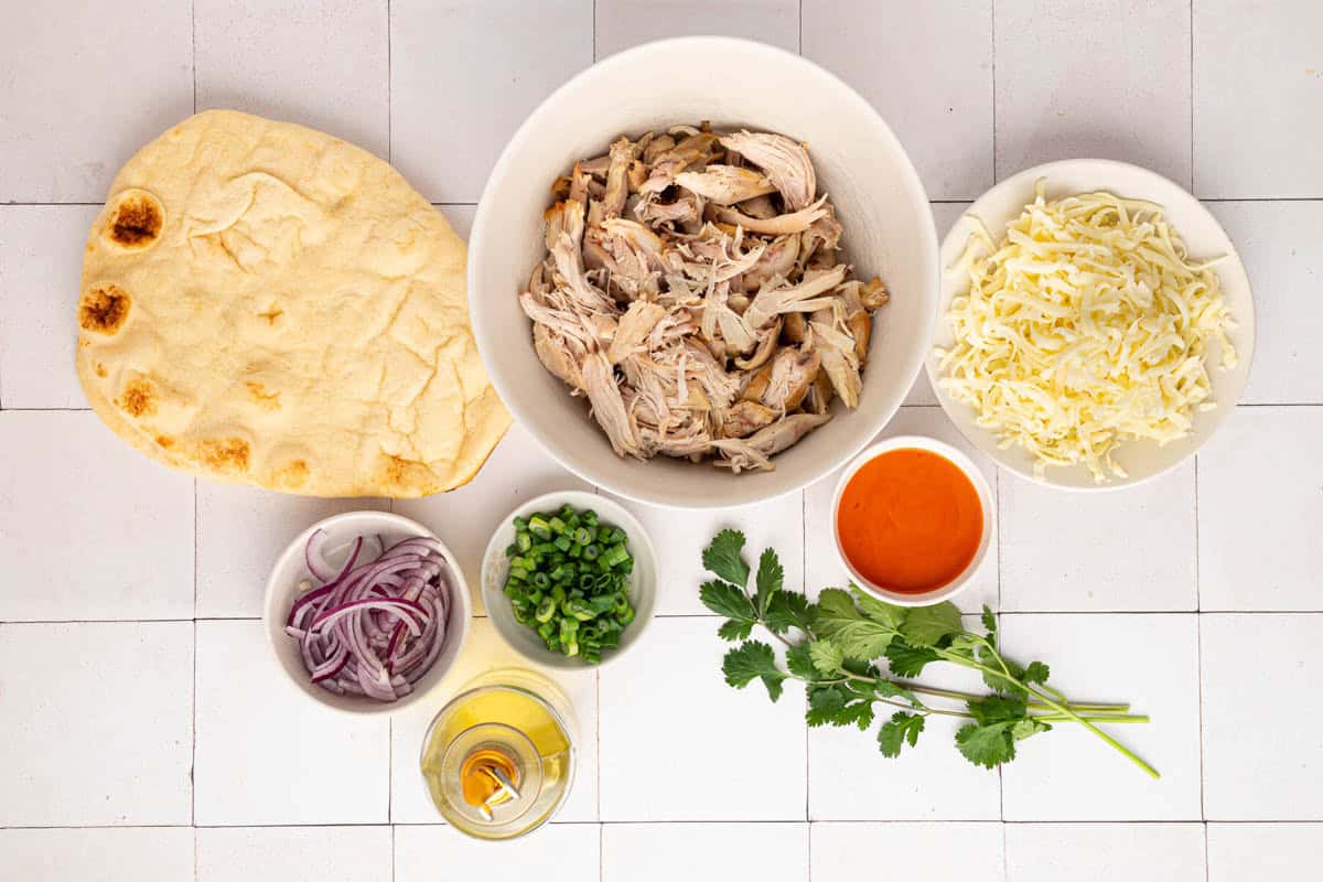 Ingredients arranged on a white tiled surface: naan bread, shredded chicken, shredded cheese, hot sauce, sliced red onion, chopped green onions, olive oil, and fresh cilantro.