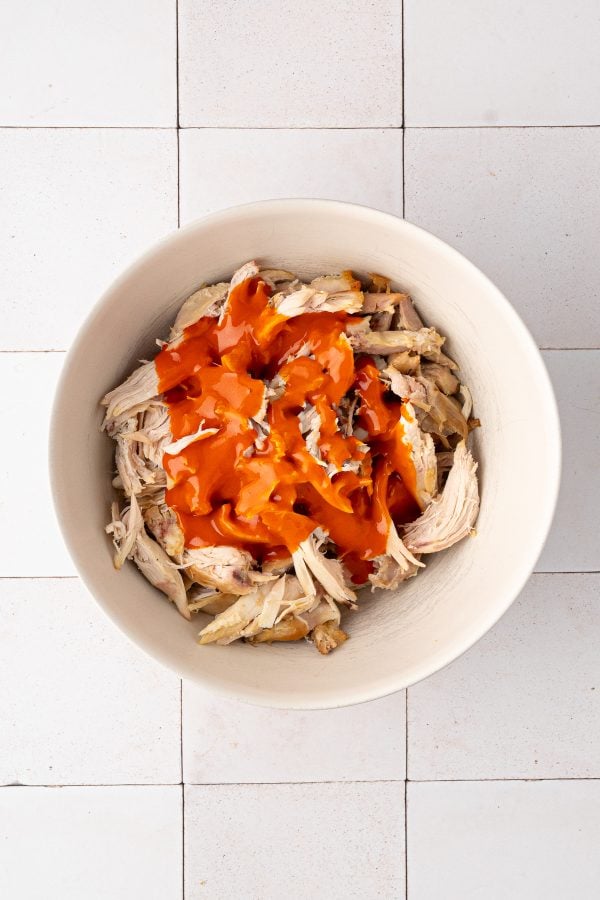 A white bowl containing shredded chicken topped with bright orange buffalo sauce, placed on a white tiled surface.