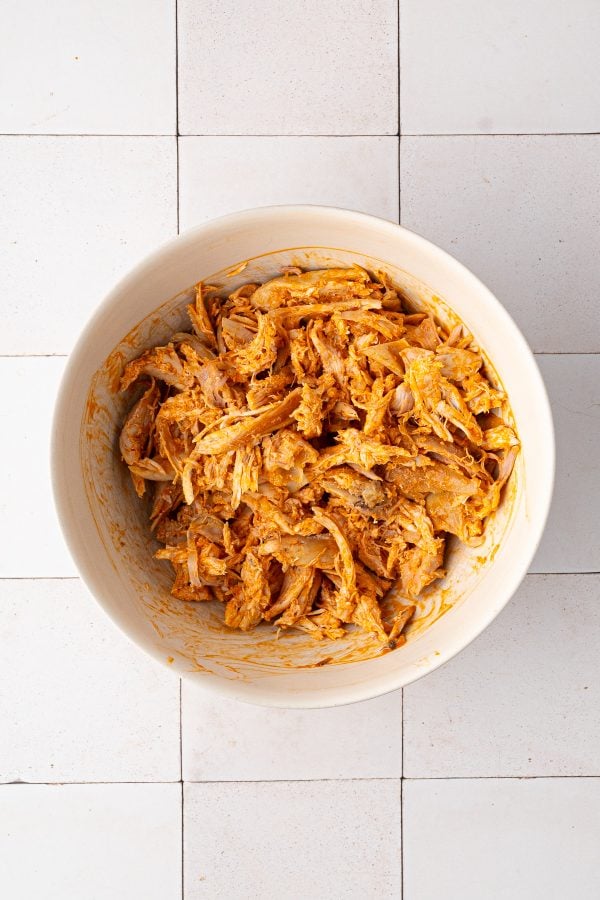 A white bowl containing shredded chicken coated in an orange-colored sauce, placed on a tiled surface.