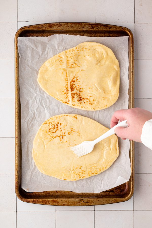 A hand using a brush to apply oil or butter to two pieces of flatbread on a parchment-lined baking sheet.