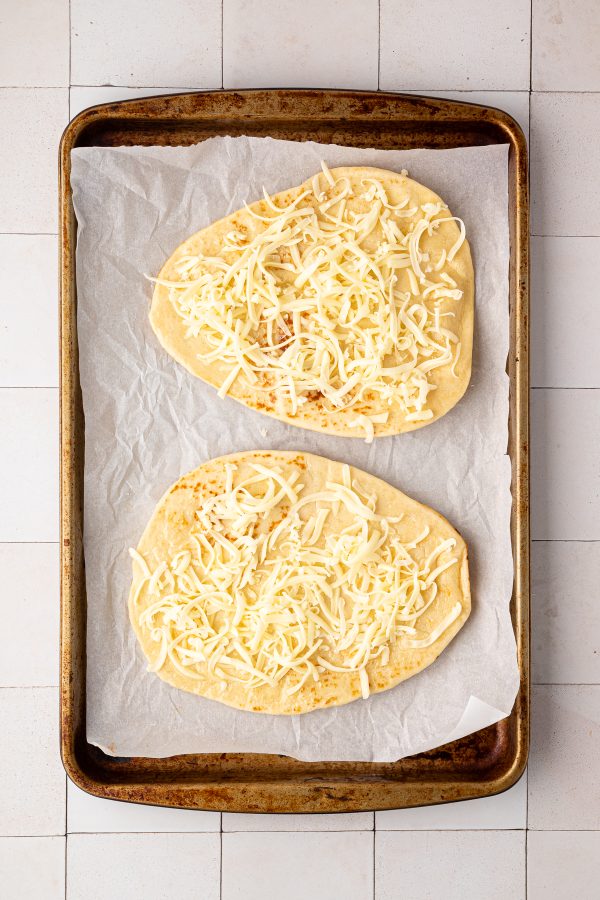 Two pieces of flatbread topped with shredded cheese on a parchment-lined baking sheet.