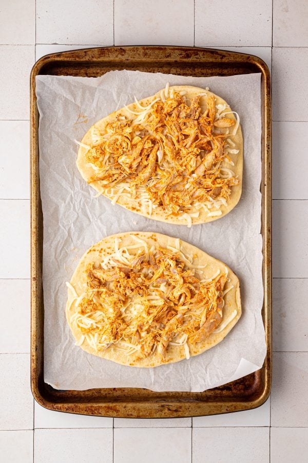 Two pieces of flatbread topped with shredded chicken and cheese on a parchment-lined baking sheet.