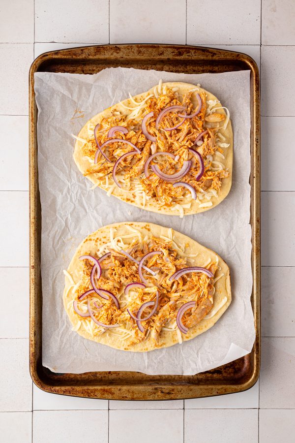 Two pieces of flatbread on a baking sheet topped with shredded cheese, seasoned chicken, and sliced red onions, ready to be baked.