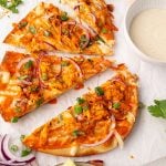 Three slices of buffalo chicken pizza topped with red onions, green onions, and cheese are arranged on parchment paper, next to a bowl of dipping sauce.