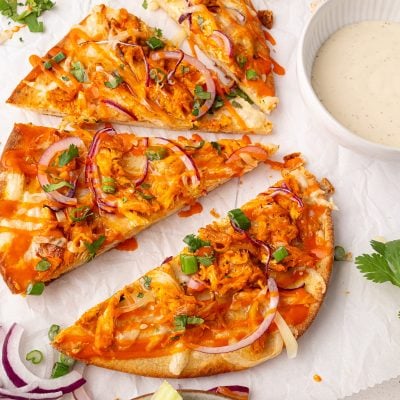 Three slices of buffalo chicken pizza topped with red onions, green onions, and cheese are arranged on parchment paper, next to a bowl of dipping sauce.