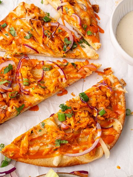 Three slices of buffalo chicken pizza topped with red onions, green onions, and cheese are arranged on parchment paper, next to a bowl of dipping sauce.