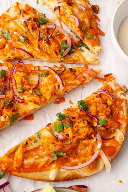 Three slices of buffalo chicken pizza topped with red onions, green onions, and cheese are arranged on parchment paper, next to a bowl of dipping sauce.