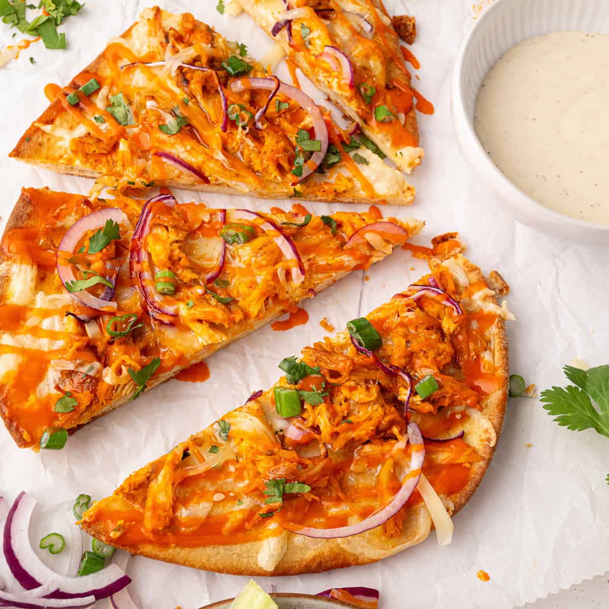 Three slices of buffalo chicken pizza topped with red onions, green onions, and cheese are arranged on parchment paper, next to a bowl of dipping sauce.