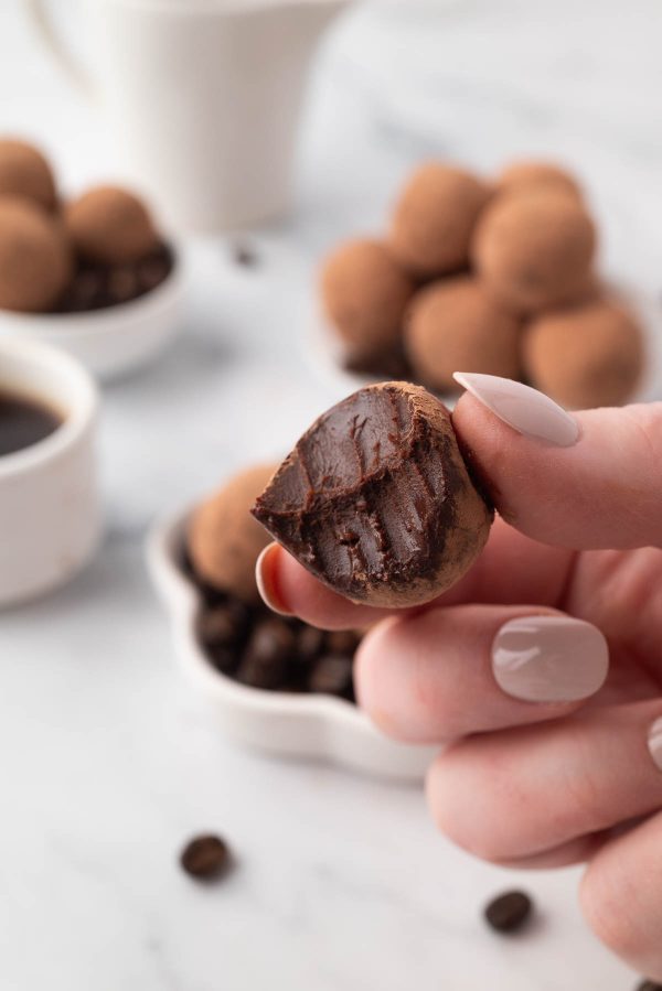 A hand holding an espresso chocolate truffle with a bite taken out, showing the smooth chocolate filling. Bowls of truffles and a cup of coffee are blurred in the background.