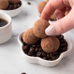 A hand picking up a cocoa-dusted chocolate truffle from a small bowl filled with coffee beans, with a cup of coffee and more truffles in the background.