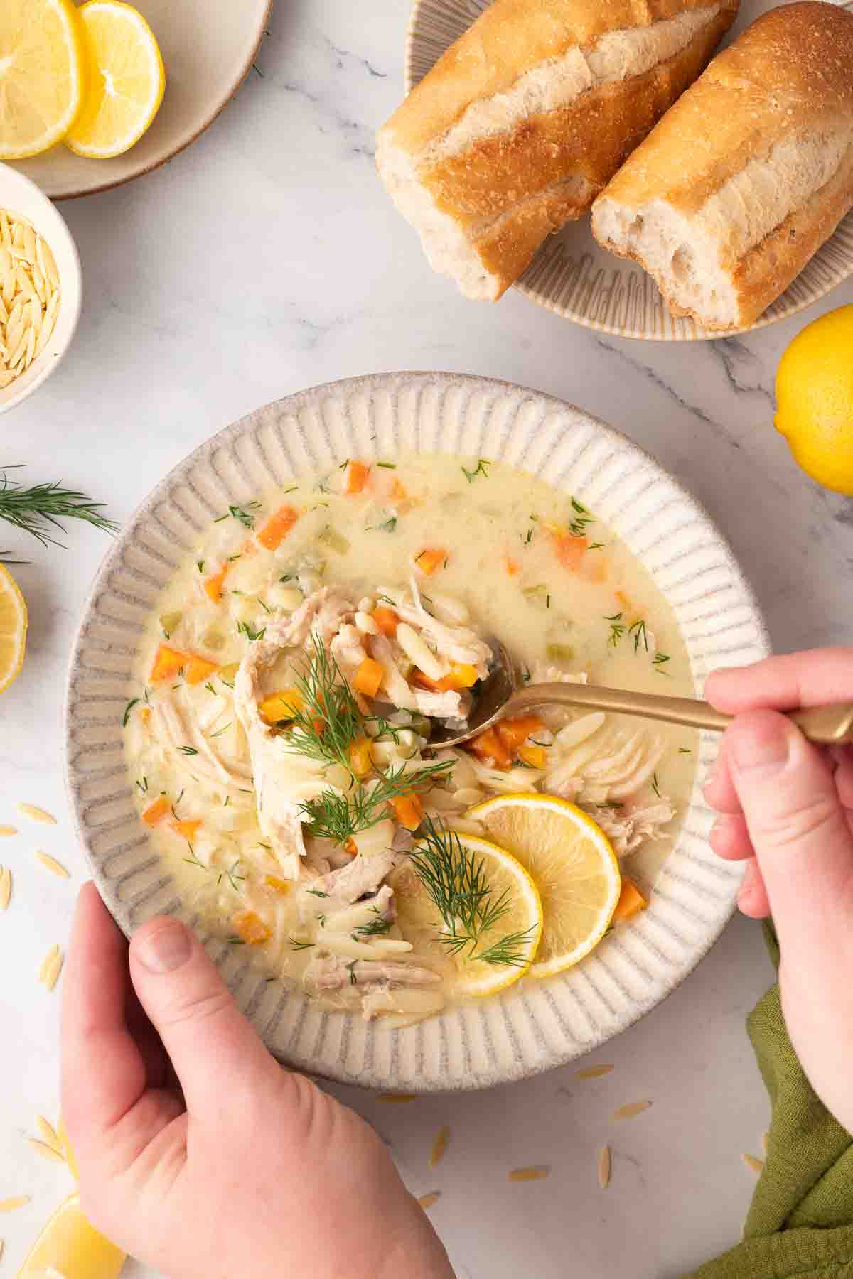 A person holds a bowl of creamy greek lemon chicken soup with orzo, garnished with lemon slices and dill, alongside a plate of sliced baguette and lemon wedges on a marble surface.