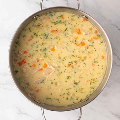 A large pot filled with creamy greek lemon chicken soup containing chunks of chicken, carrots, and fresh herbs, viewed from above on a marble surface.