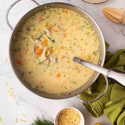 A pot of greek lemon chicken soup garnished with dill, with a ladle inside. Surrounding the pot are fresh dill, a green towel, lemon slices, and uncooked orzo.