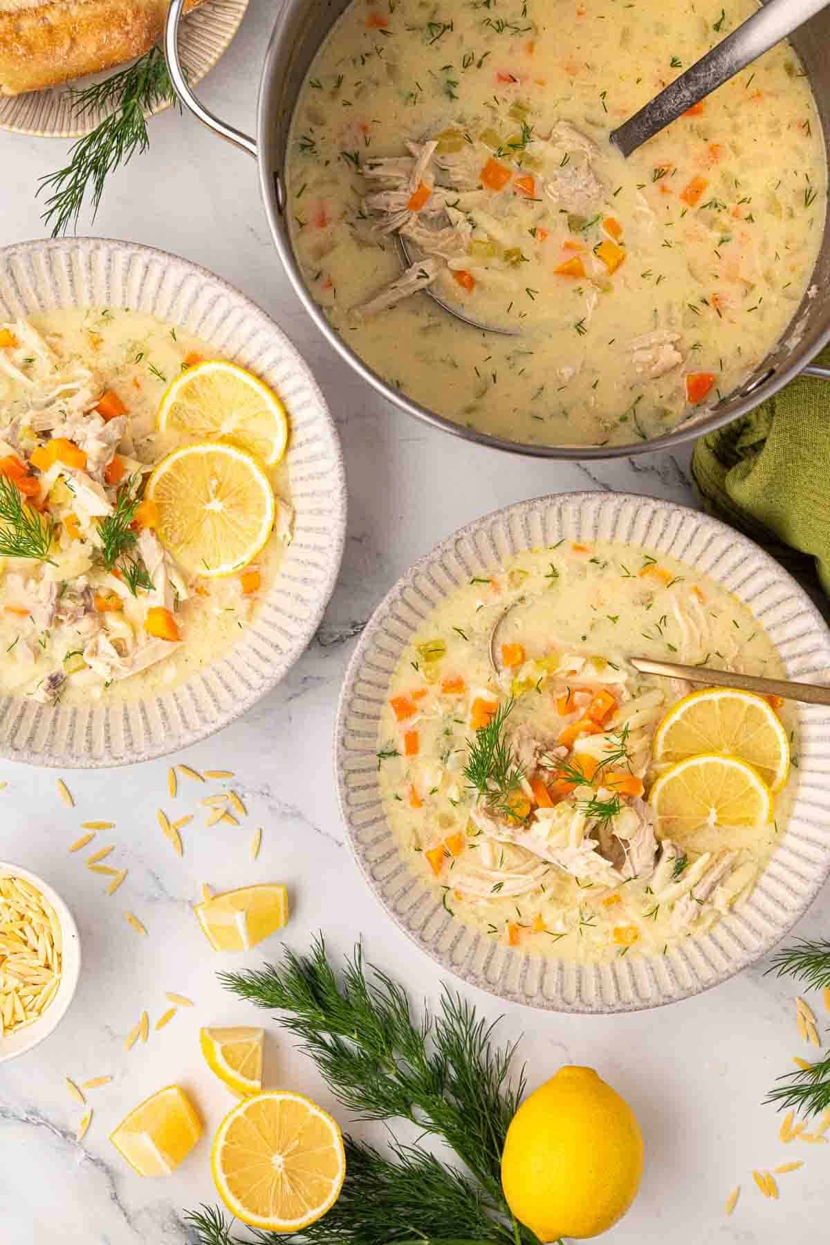 Two bowls and a pot of lemon chicken soup with carrots and dill, garnished with lemon slices and fresh dill, surrounded by lemon wedges, orzo, and bread.