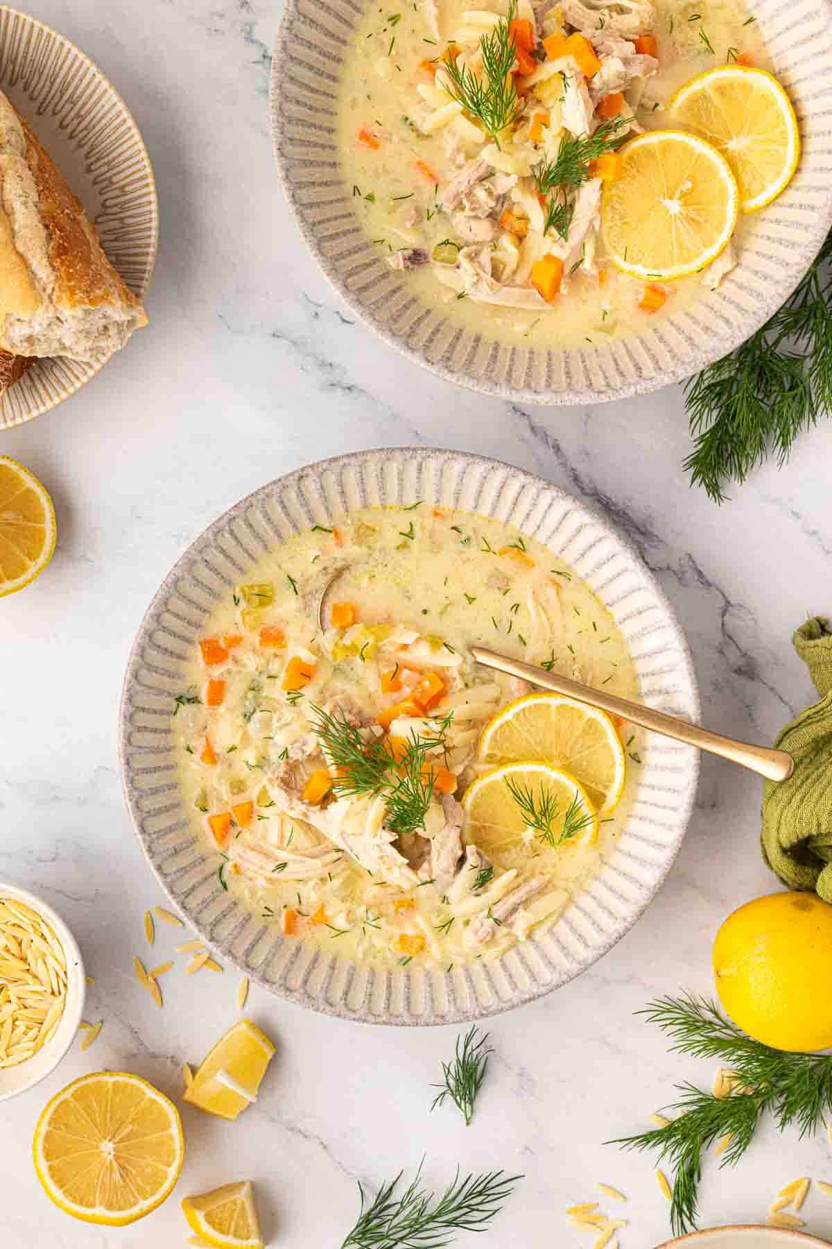 Two bowls of creamy greek lemon chicken soup garnished with fresh dill and lemon slices, surrounded by bread, lemon wedges, fresh dill, and uncooked orzo on a marble surface.