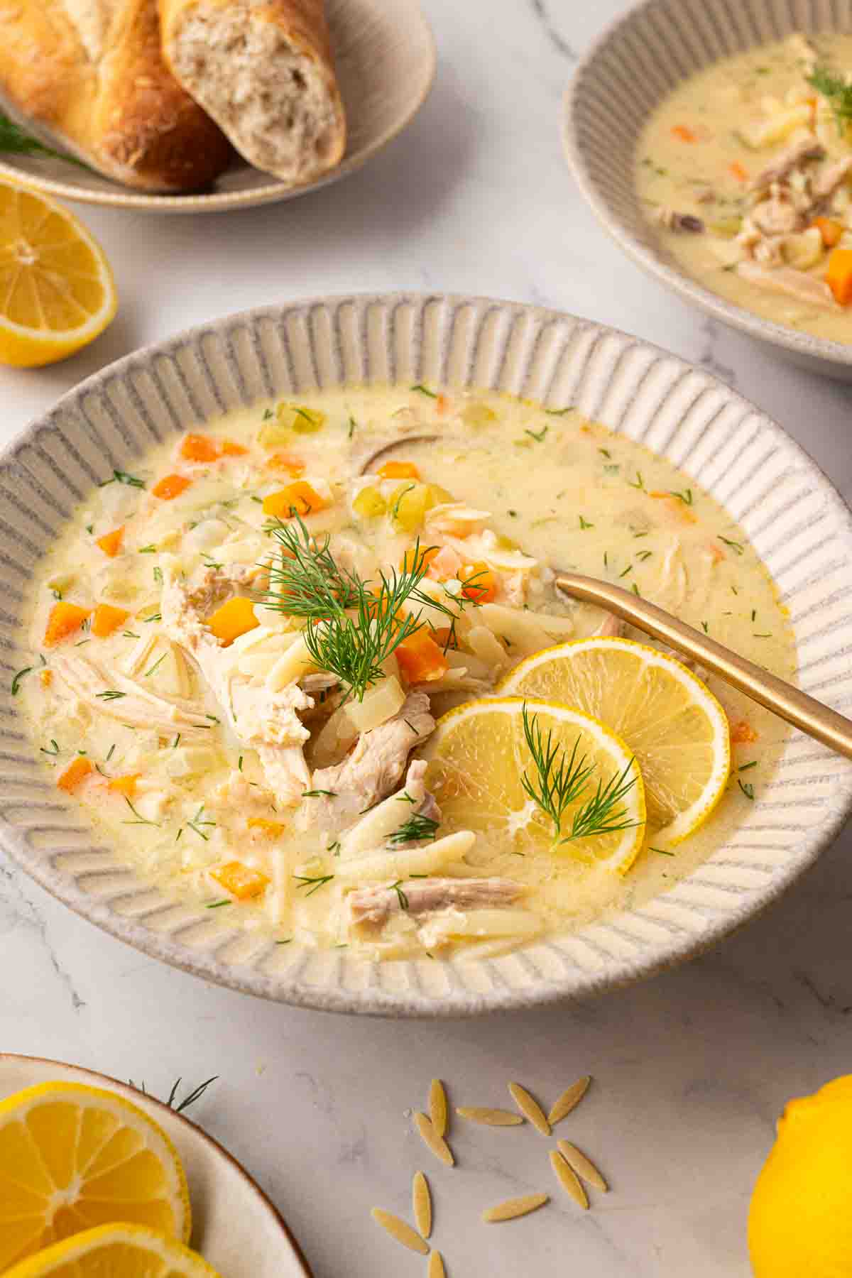 A bowl of creamy chicken soup with shredded chicken, orzo, diced carrots, fresh dill, and two lemon slices, served with a spoon on a white plate.
