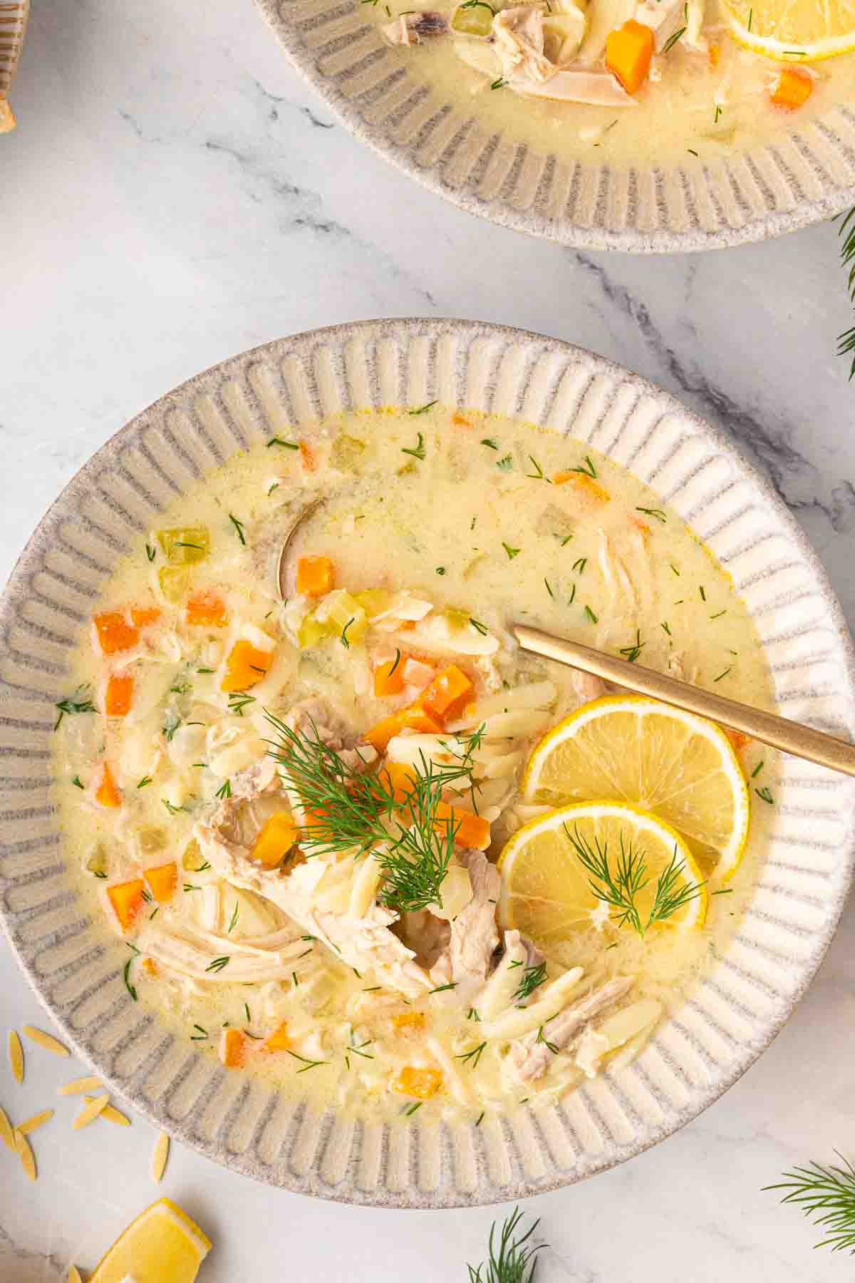 A bowl of creamy chicken soup with shredded chicken, orzo, diced carrots, fresh dill, and two lemon slices, served with a spoon on a white plate.