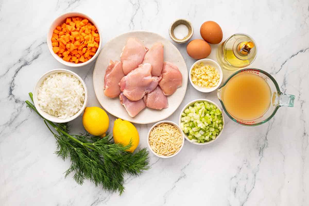 Ingredients for a recipe arranged on a marble surface, including chicken, eggs, broth, rice, carrots, celery, garlic, dill, lemon, cheese, oil, and salt.