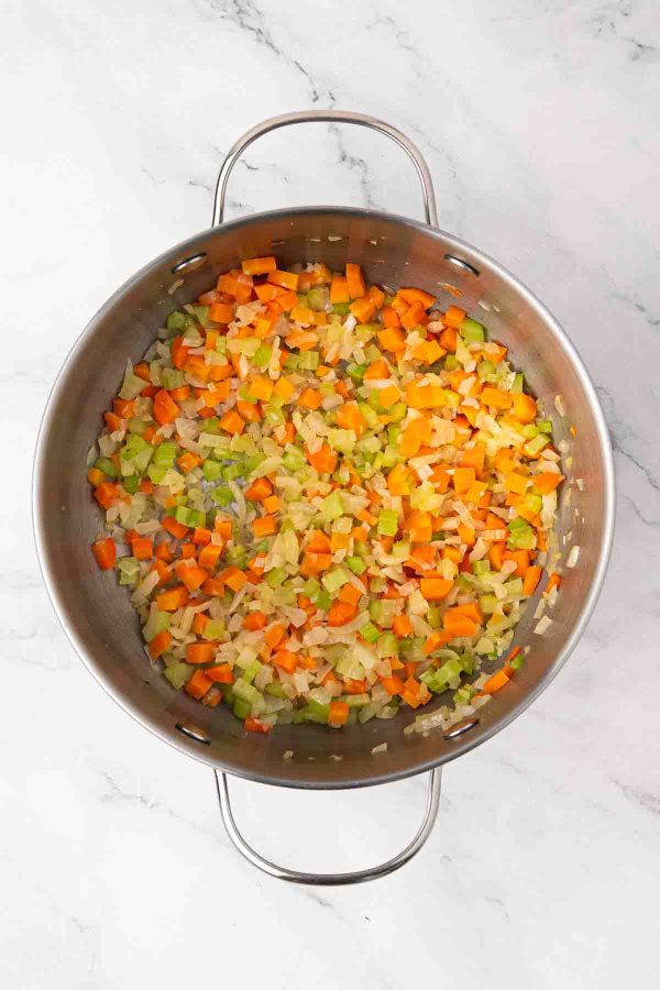 Chopped carrots, celery, and onions saut&eacute;ing in a large stainless steel pan on a marble countertop.