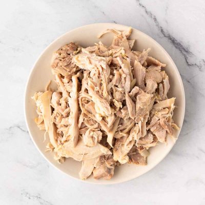 A white plate filled with shredded cooked chicken sits on a light marble surface.