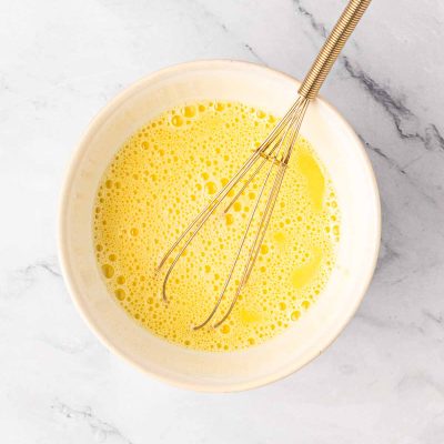 A metal whisk rests in a white bowl filled with beaten eggs on a marble countertop.