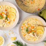 A bowl of creamy chicken soup with carrots, orzo, lemon slices, and fresh dill, served with a spoon on a marble surface.