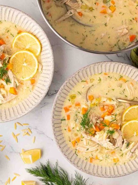 A bowl of creamy chicken soup with carrots, orzo, lemon slices, and fresh dill, served with a spoon on a marble surface.