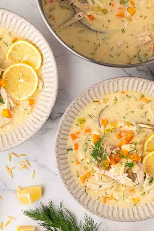 A bowl of creamy chicken soup with carrots, orzo, lemon slices, and fresh dill, served with a spoon on a marble surface.