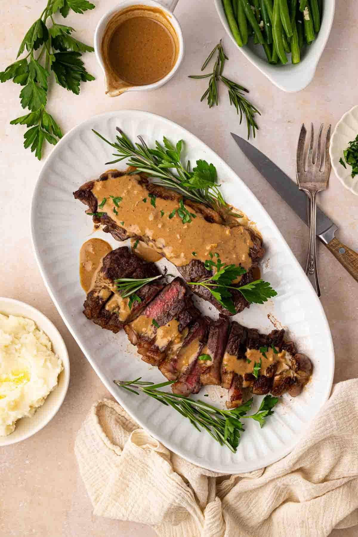 Sliced steak topped with brown sauce and herbs on a white platter, served with mashed potatoes, green beans, and garnished with rosemary and parsley.