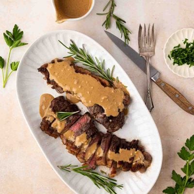 Sliced steak topped with creamy sauce and garnished with rosemary, served on a white platter with a sauce pitcher, herbs, knife, and fork nearby.
