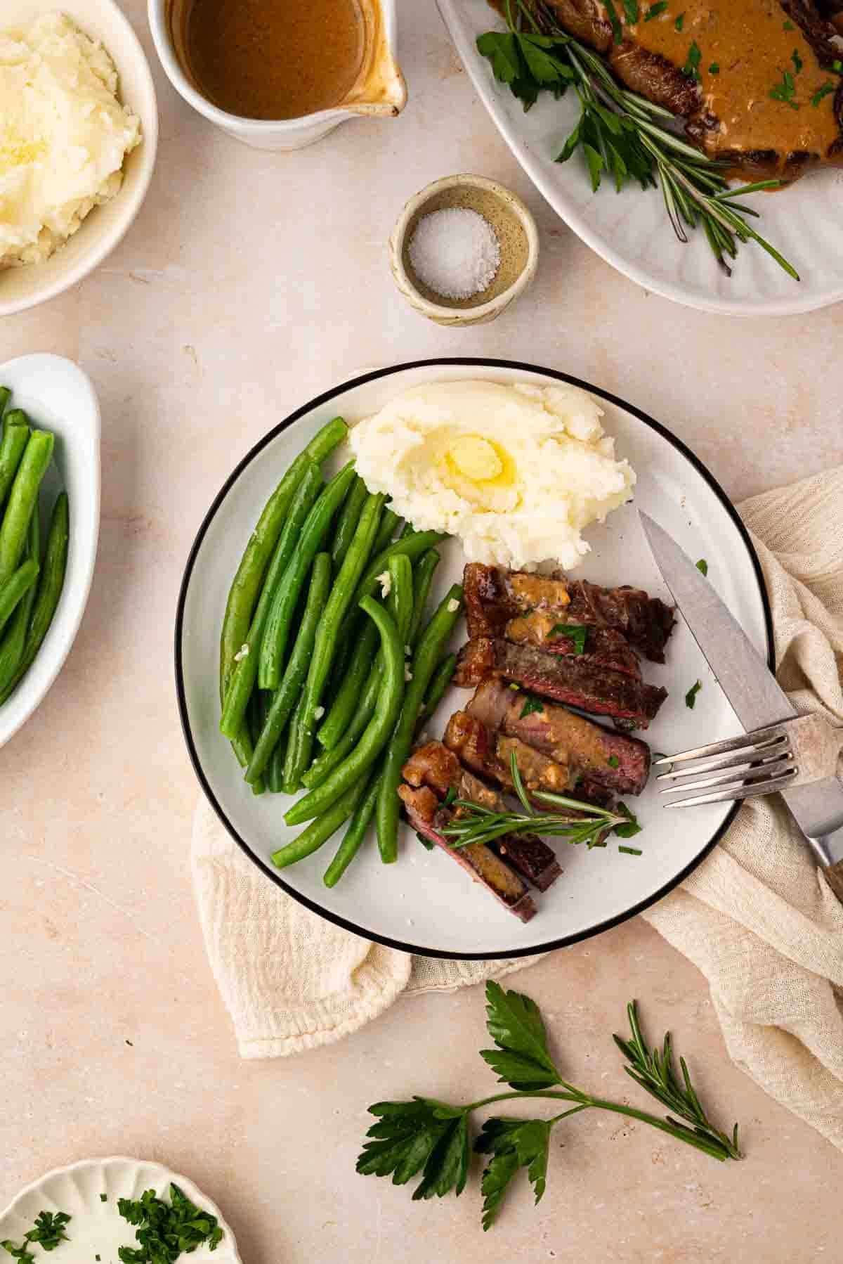 A plate with sliced steak topped with gravy, mashed potatoes with butter, and green beans, garnished with parsley. A fork and knife rest on the plate.