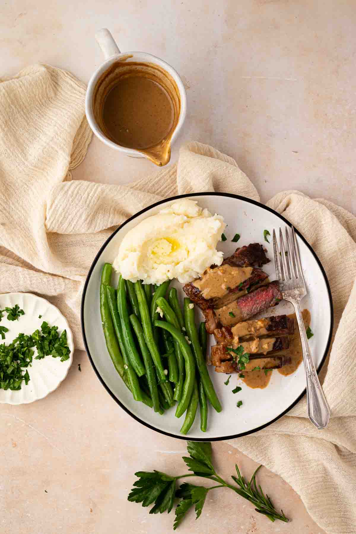 A plate with mashed potatoes, green beans, and sliced steak topped with gravy, accompanied by a fork; gravy boat and chopped herbs on the side.