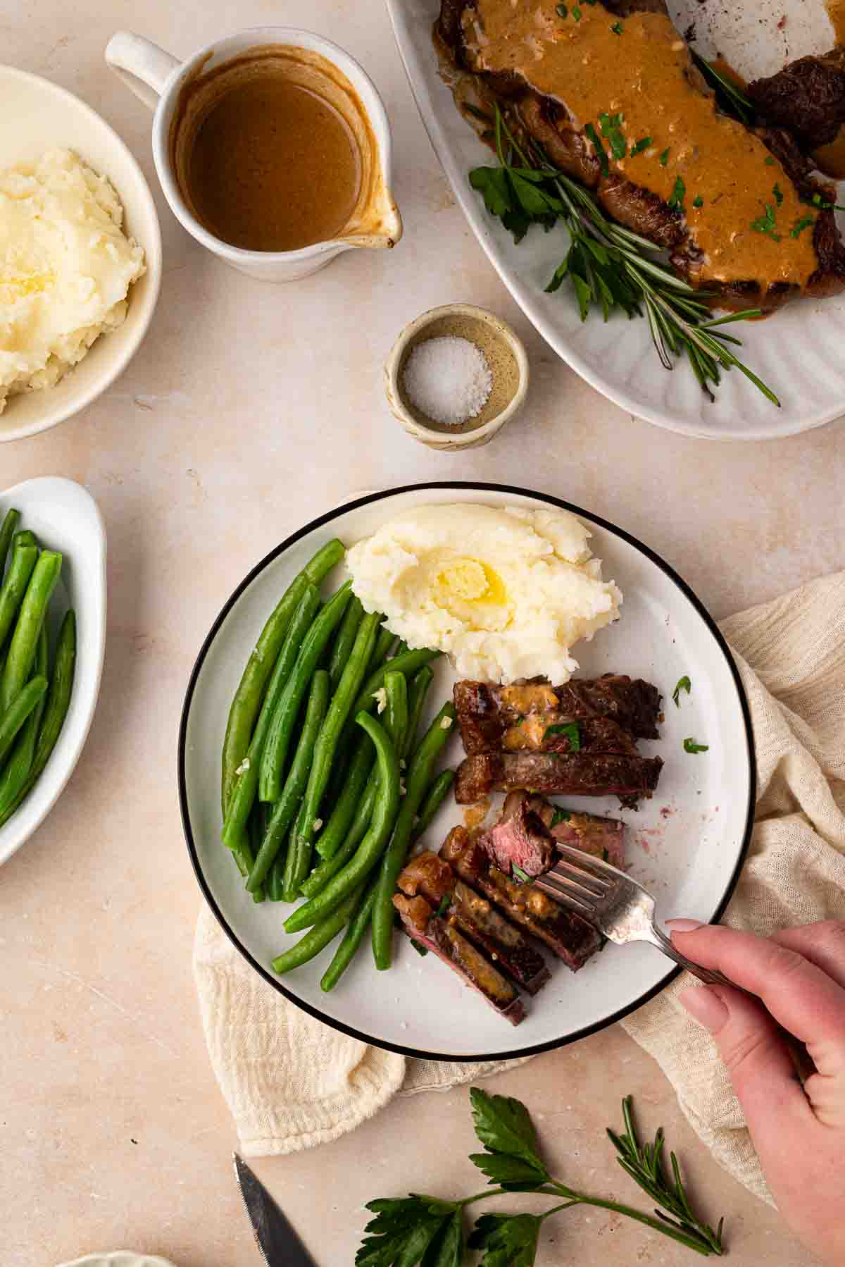 A plate with sliced steak, mashed potatoes topped with butter, and green beans. Nearby are a gravy jug, a dish of mashed potatoes, a platter of steak, and a bowl of green beans.