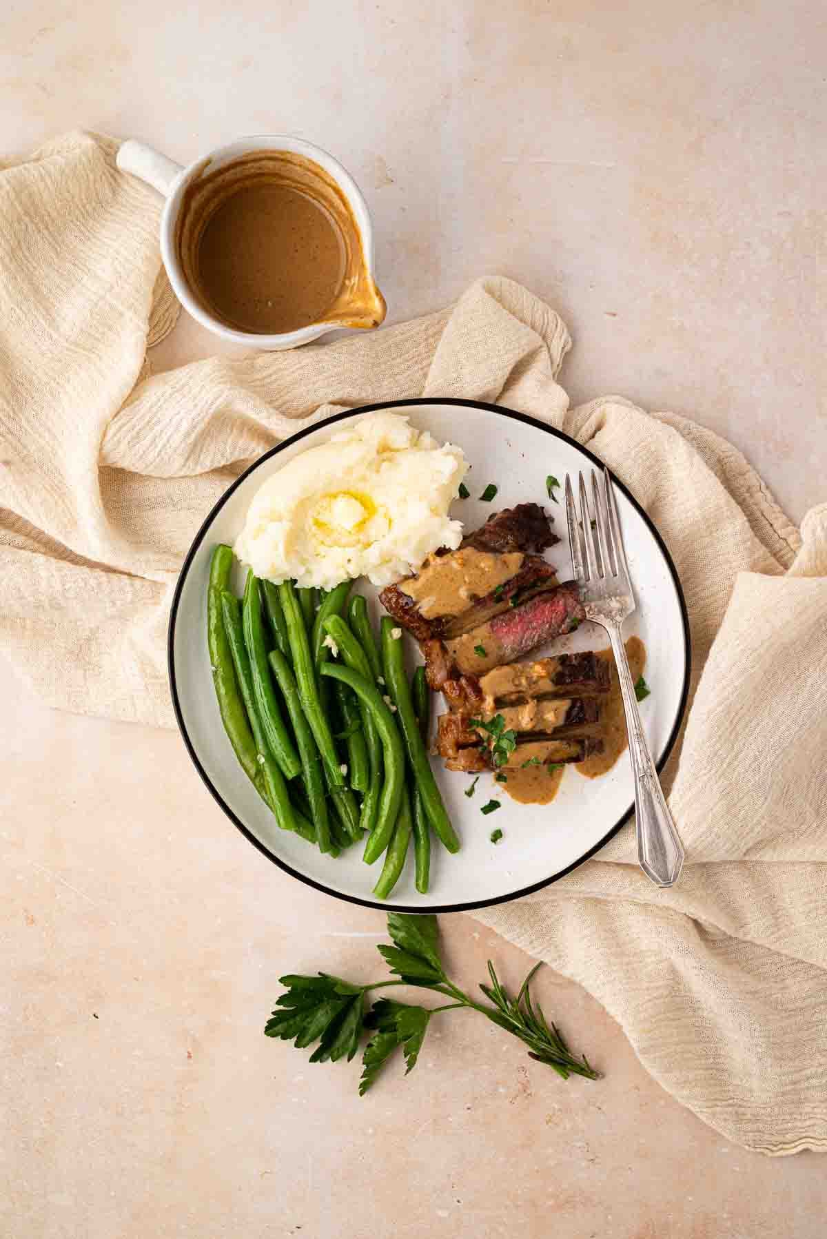 A plate with mashed potatoes, green beans, and sliced steak topped with gravy, served with a fork and knife. A small pitcher of gravy and herbs are nearby on a beige cloth.