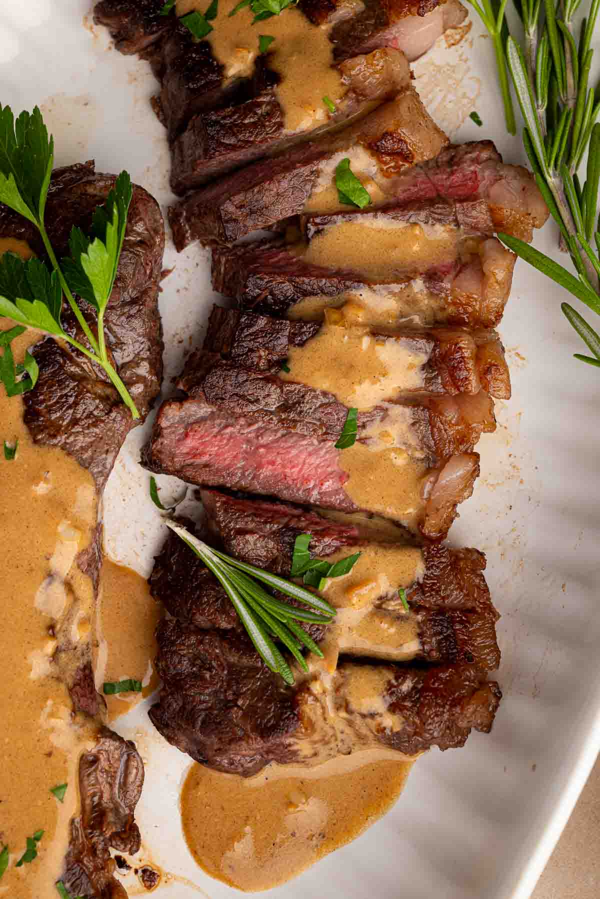 Sliced steak topped with creamy brown sauce, garnished with fresh herbs, served on a white plate.