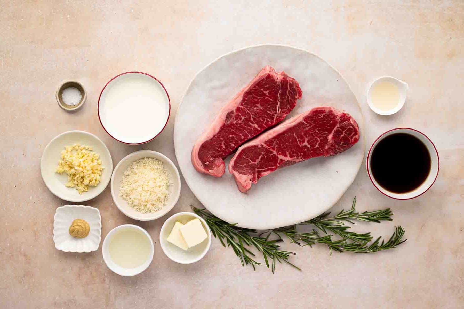 Two raw steaks on a white plate surrounded by small bowls containing cream, soy sauce, butter, oil, minced garlic, grated cheese, mustard, salt, and sprigs of fresh rosemary.