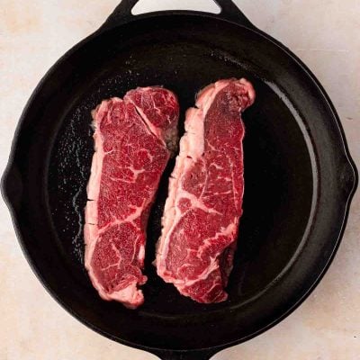 Two raw strip steaks are placed side by side in a black cast iron skillet on a light-colored surface.