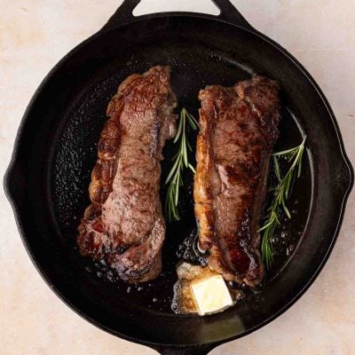 Two cooked steaks with sprigs of rosemary and a pat of melting butter in a black cast iron skillet.