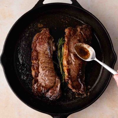 Two cooked steaks in a cast iron skillet with a sprig of rosemary, as a hand spoons sauce or juices over one of the steaks.