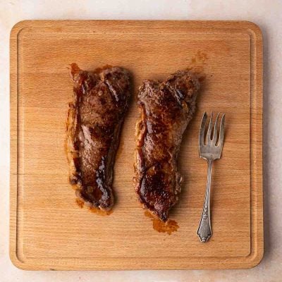 Two cooked steak strips and a meat fork are placed on a wooden cutting board, set on a light-colored surface.